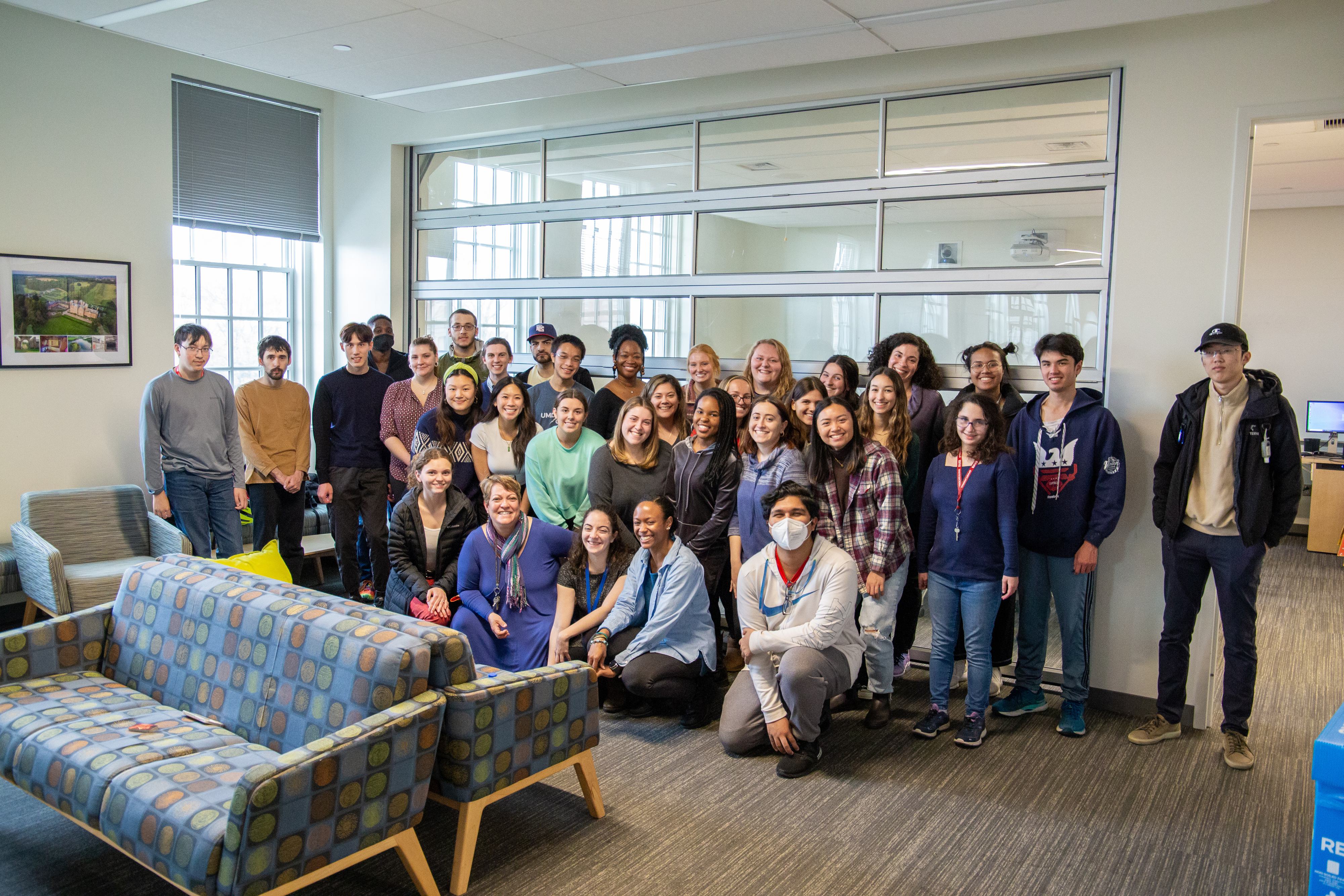 Dozens of people pose for a photo while standing and crouching in a room. Sofas are in the foreground and windows are in the background.