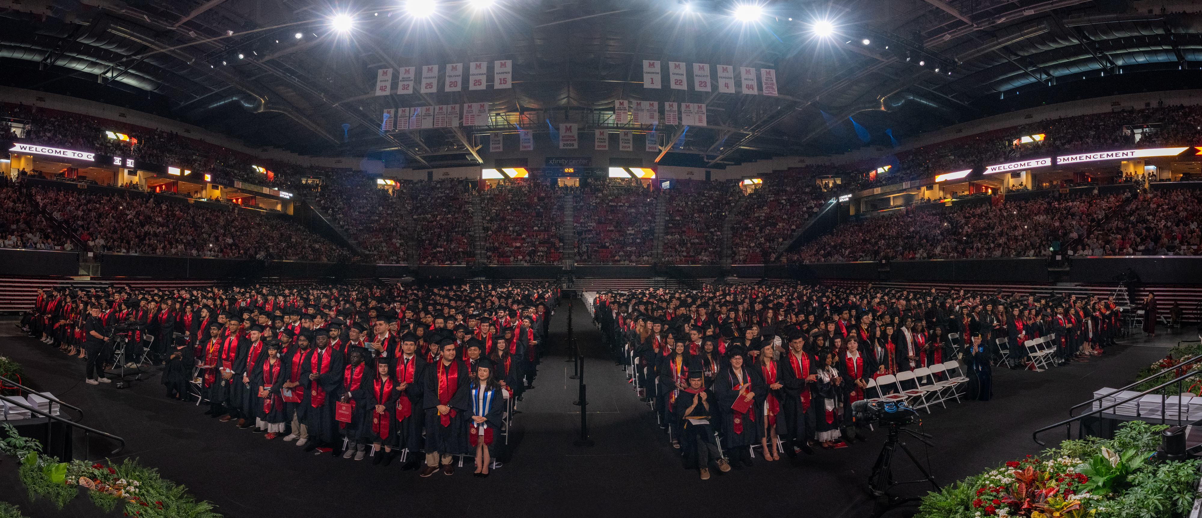 A wide shot of a full arena with graduating students wearing black and red regalia.