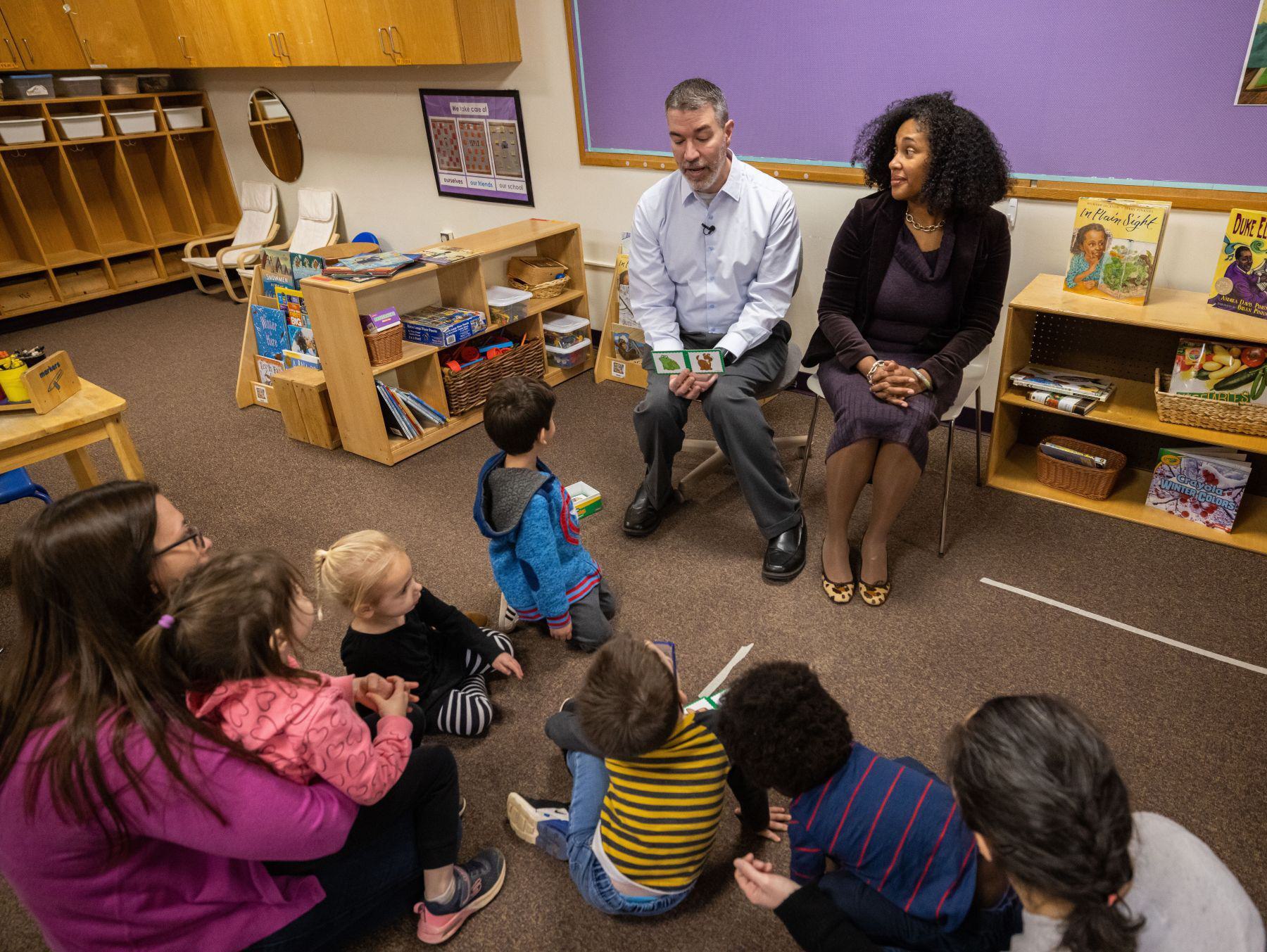 Two people sit in chairs in a classroom as children on the carpeted floor look up at them intently. There are cubbies and a purple bulletin board in the background.