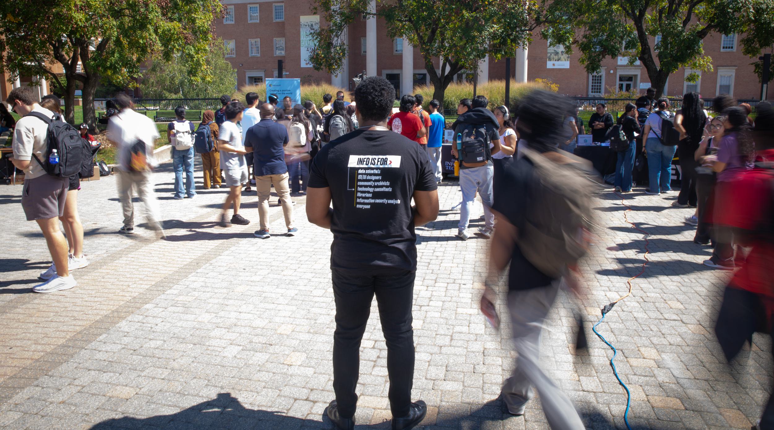 A man standing in a the middle of a crowded square, wearing a black shirt, as people walk past him.