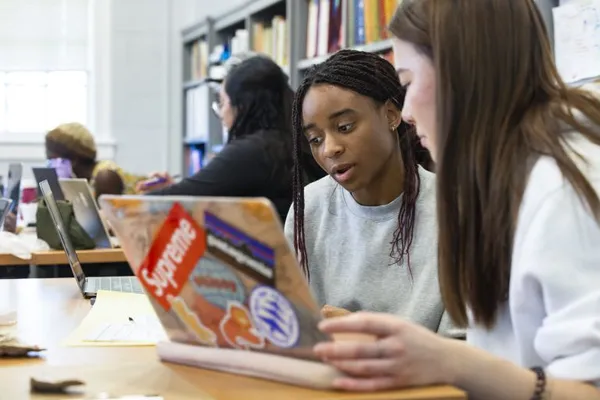 Two people sitting together, looking at a laptop and some pieces of paper as though they are studying. There are books on a shelf in the background.