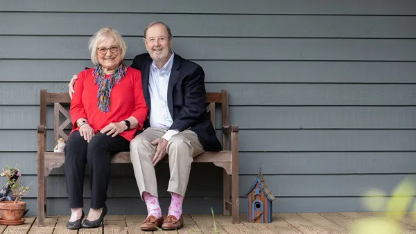 A man and woman sit on a bench in front of gray siding. The man has his right arm around the shoulder of the woman.