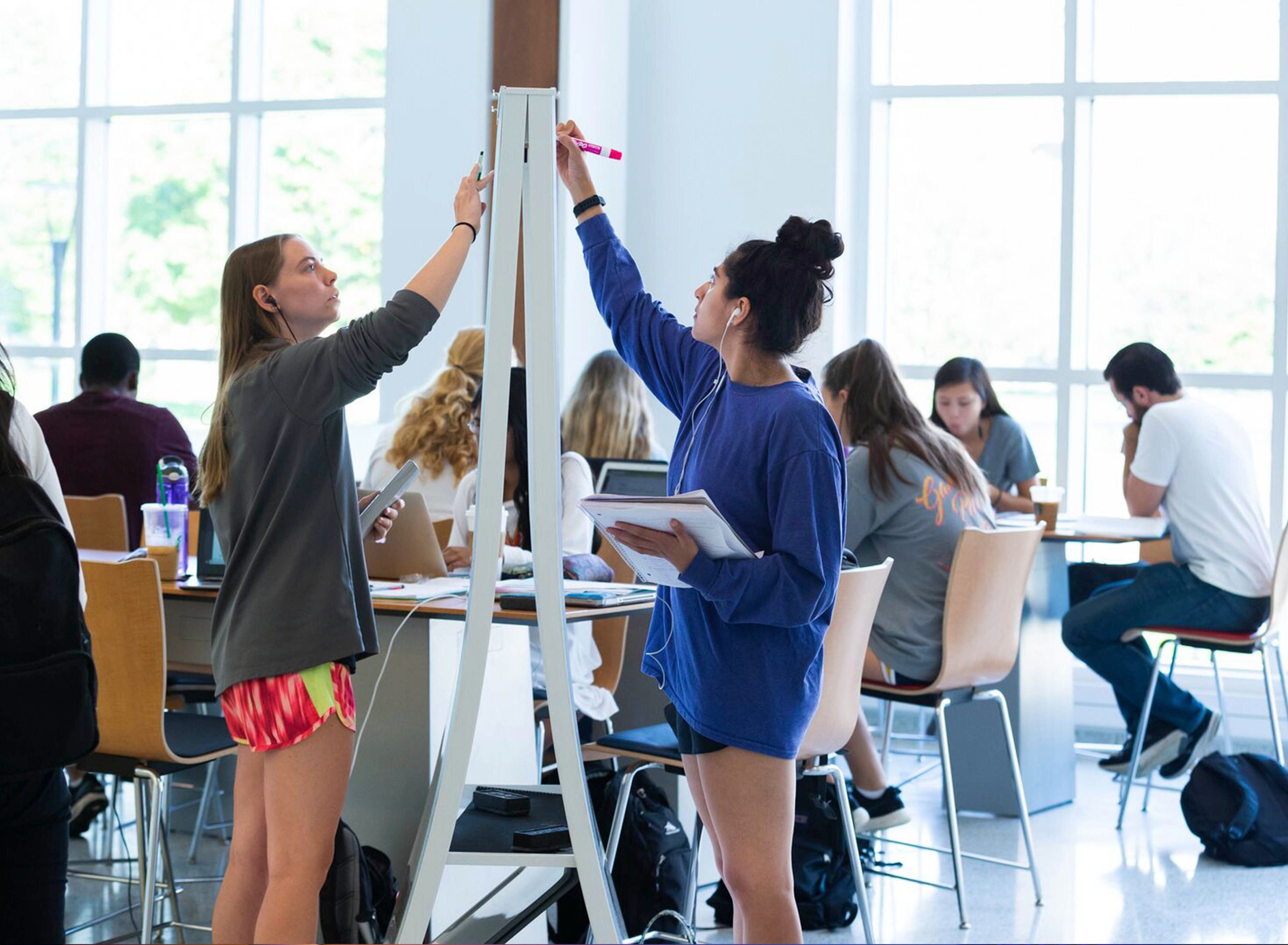 Two students standing at a tall whiteboard, writing with markers on opposite sides, in a bright study space where other classmates sit at tables and work in the background