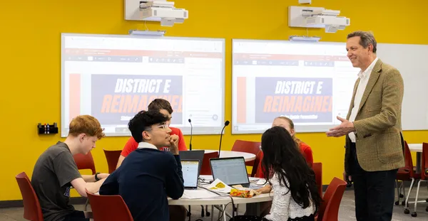 A person stands in front of a group of students in a room with yellow walls.