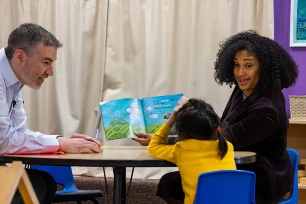 Two adults and a child reading a picture book in a classroom environment.