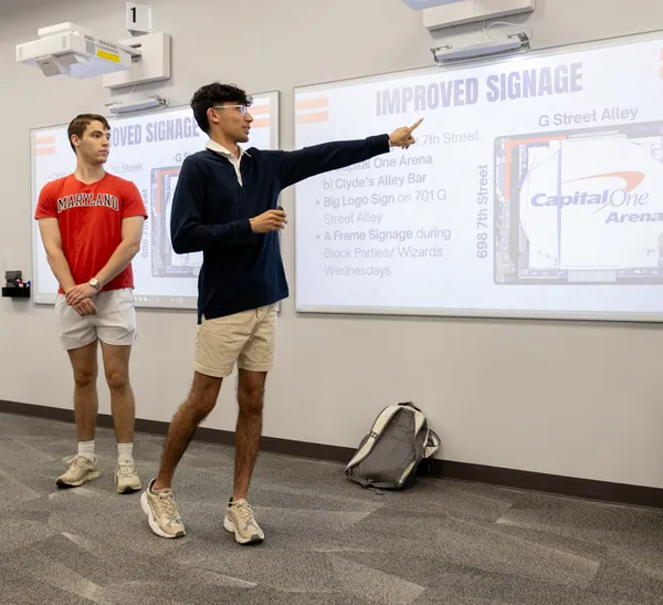 Two students stand in front of a projection screen, pointing to the images on the screen.