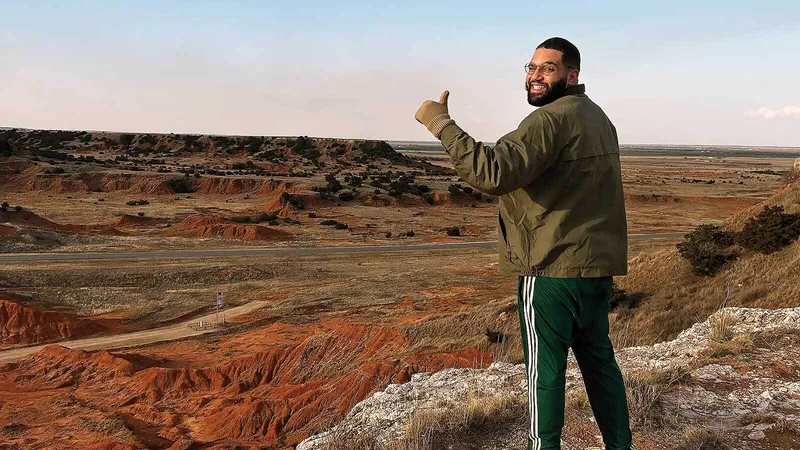 A man standing over a vast landscape of brown, orange, and gray colors looks back over his shoulder and gives a thumbs up.