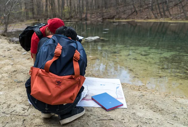 Two people crouch next to a small body of water, observing a piece of paper with some architectural design that is out of focus on it.