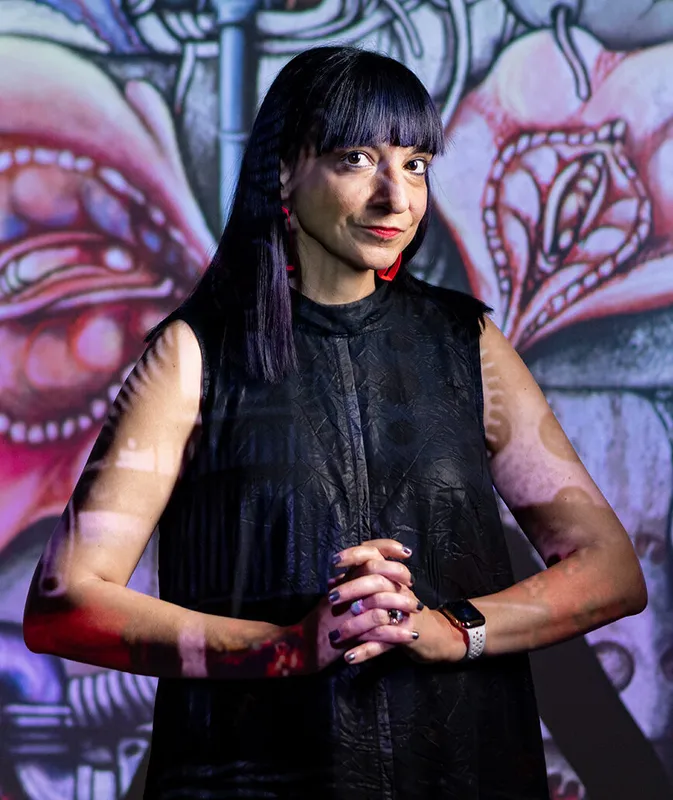 A person folds their hands in front of them, wearing a black dress, standing in front of a wall with a painting of flowers.