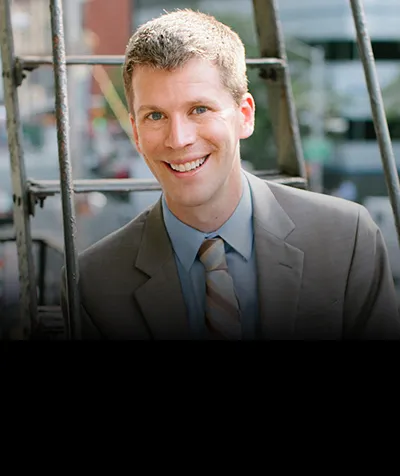 A man smiles, wearing a light gray suit with a light blue shirt and a striped tie. There are steps for a fire escape behind him, with a city street beyond.