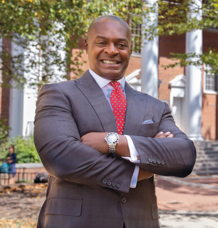 A man stands with his arms folded across his chest, wearing a red tie and dark gray suit. A building and trees are behind him.