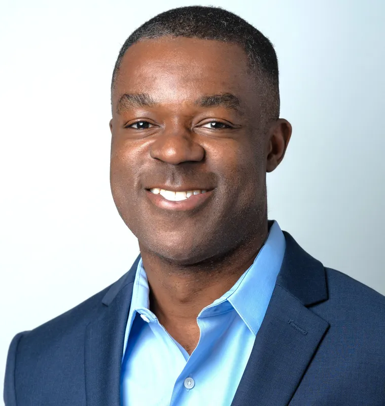 A man smiles, wearing a light blue dress shirt under a dark blue suit jacket.