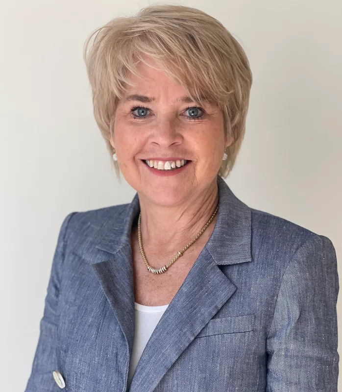 A woman smiles, wearing a necklace, a white top, and a blue-gray suit jacket.