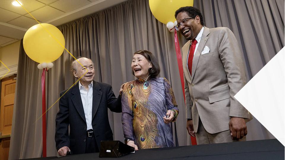 An older man in a dark suit, a woman in a colorful patterned dress, and President Pines stand behind a table in front of gray curtains, smiling as two large gold balloons and red ribbons float above them