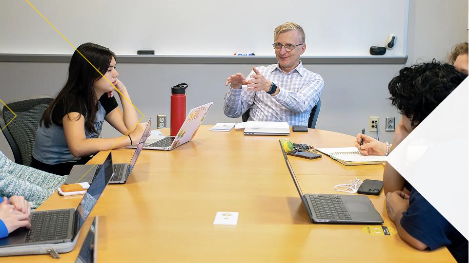 An instructor seated at the head of a wooden conference table gestures with his hands while students around the table take notes and work on laptops, with a whiteboard visible on the wall behind them