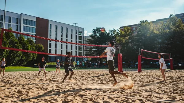 People are playing beach volleyball on courts with sand and red nets. It is a sunny day with blue sky.