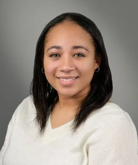 A person with shoulder length black hair smiles while wearing a white sweater.