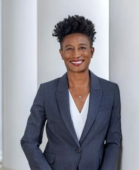 A person wearing a white shirt and dark gray suit smiles while leaning against a white column on a building.