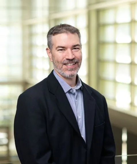 A person wearing a light blue shirt and black sports coat smiles while standing in front of a bay of windows.