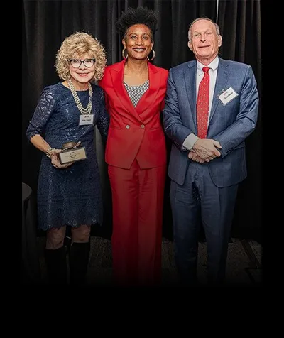 Three people stand side by side smiling. The woman on the left is holding a small trophy of a turtle.