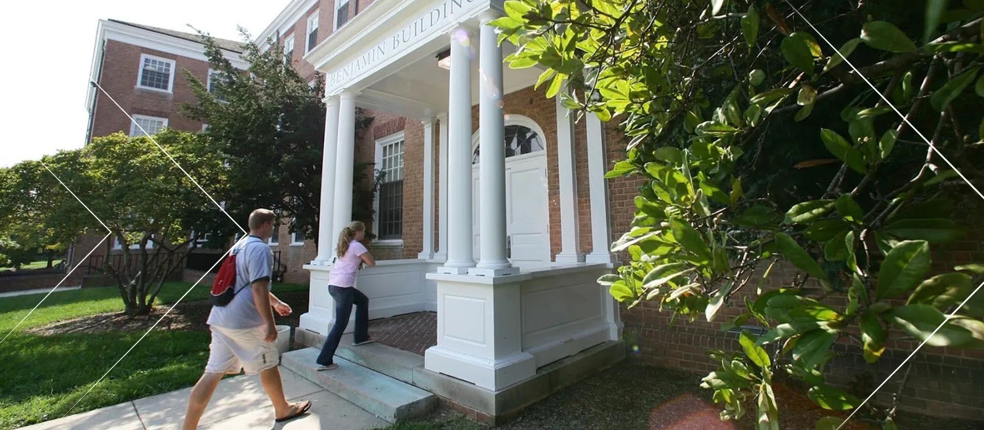 Two people are walking on a sidewalk into the entrance of a building marked "Benjamin Building," with small trees on either side.