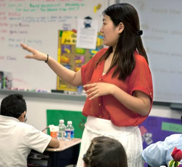 A woman wearing a red shirt gestures with her hands while standing in front of a classroom of students.