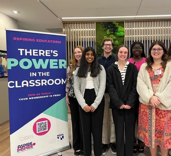 Six people stand with their hands folded in front of them, posing for a photo while standing next to a tall banner that reads, "There's Power in the Classroom."