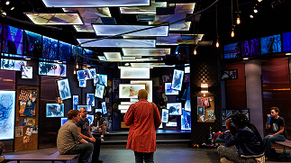 A dimly lit gallery-style space in the College of Arts and Humanities, with dozens of wall-mounted digital screens and floating illuminated panels overhead, and several people seated on benches observing the multimedia installation.