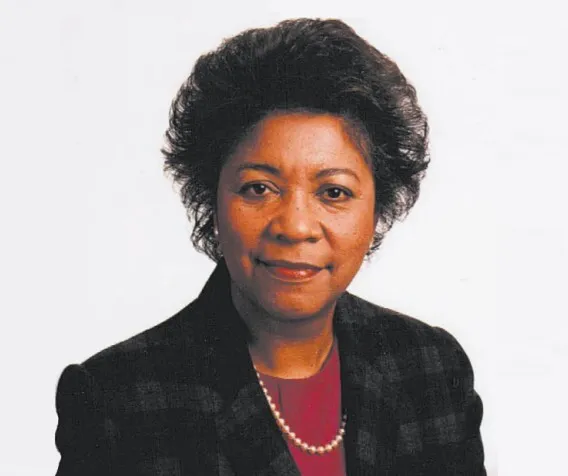 A woman wearing a red shirt, a black blazer, and pearls.