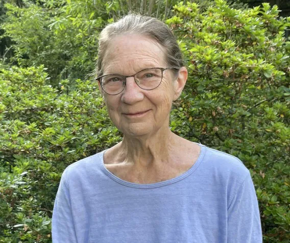 A woman wearing glasses and a blue shirt smiles, there is a green tree in the background.