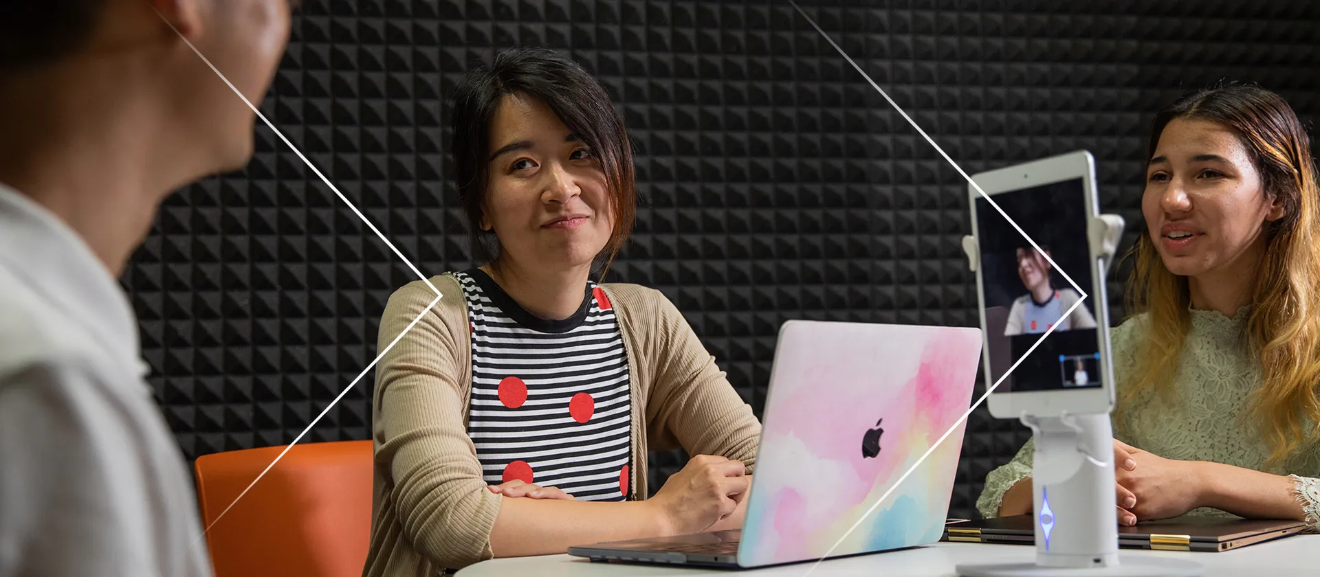 A woman, center, smiles as she looks at a person on the left, a computer is in front of her, and another person is to the right.
