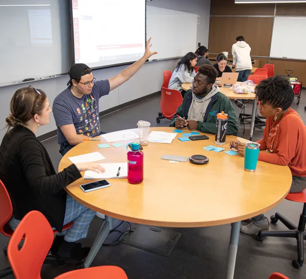 A group of students engaging with one another while sitting around a circular table. One is wearing a backward hat and gesturing with his left hand as the other listen to what he is saying.