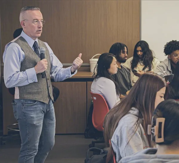 A person wearing a blue shirt, blue tie, and gray vest holds a microphone as he addresses people in the room.