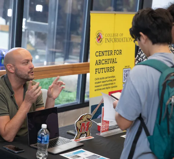A person wearing a green shirt sits a a table with a black tablecloth, speaking to two people in front of him on the other side of the table. There is a laptop, a cardboard cut out of a turtle, and other items on the table.