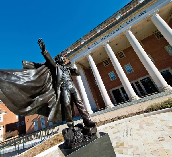 A statue of a man holding up his right hand, with his mouth open as though shouting, and his long coat billowing in the wind. In the background, a building labeled "R. Lee Hornbake Library."