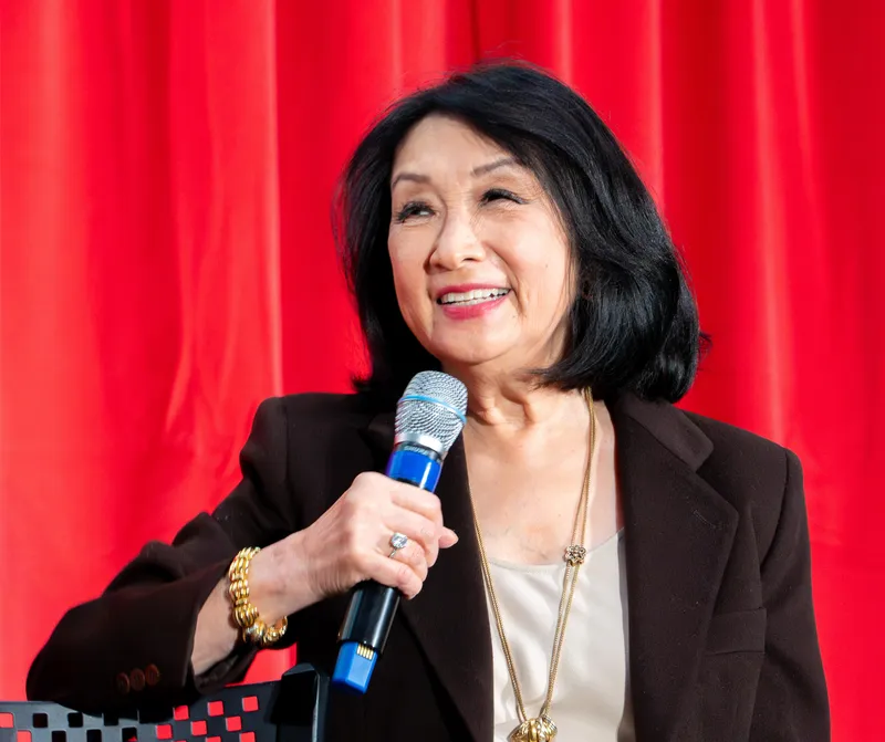 A woman with shoulder length black hair, holding a microphone in her right hand, smiling, while positioned in front of a red curtain.