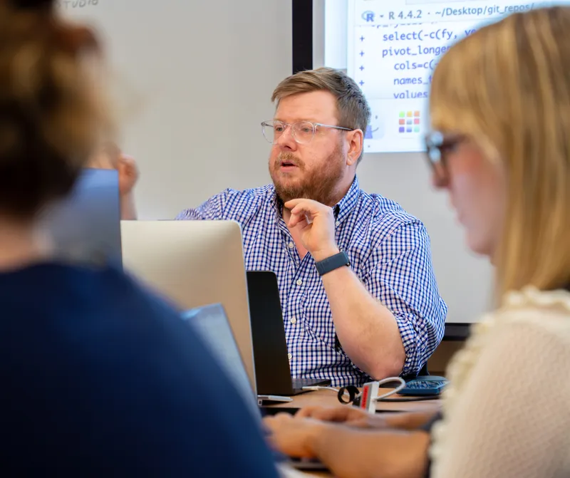 A man wearing glasses and a blue button up shirt is speaking, other people are in the room with him, and computers can be seen on the table in front of him.