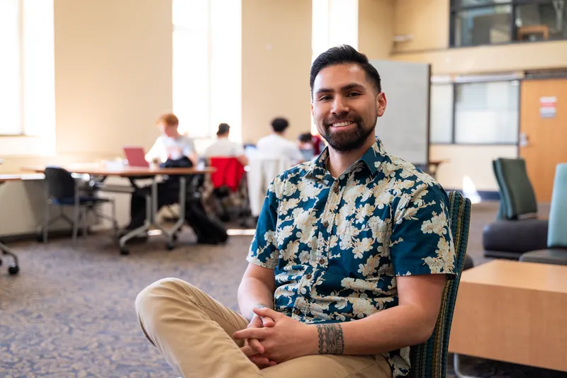 A person sits with one leg crossed over the other, smiling from inside a library study space with people at tables in the background.