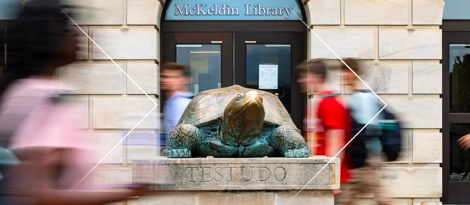 Students walk past a turtle statue, which has the name "Testudo" written on it, with the words "McKeldin Library" on the door of the building in the background.