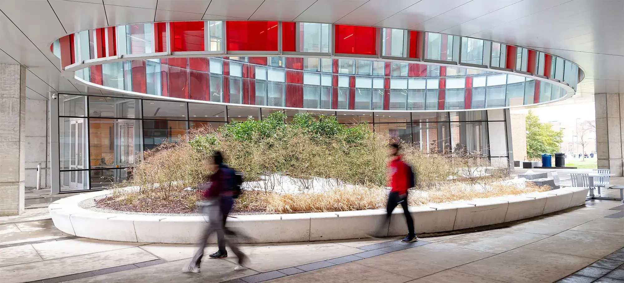 Two blurred pedestrians walk past on the tiled floor in front of the Physical Sciences building at UMD featuring tall windows and red panels.
