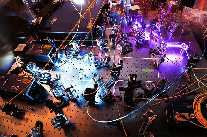 An optical table in a scientific laboratory covered with metal mounts, mirrors, lenses, and cables, with purple and blue laser beams scattering across the tabletop and illuminating the equipment.