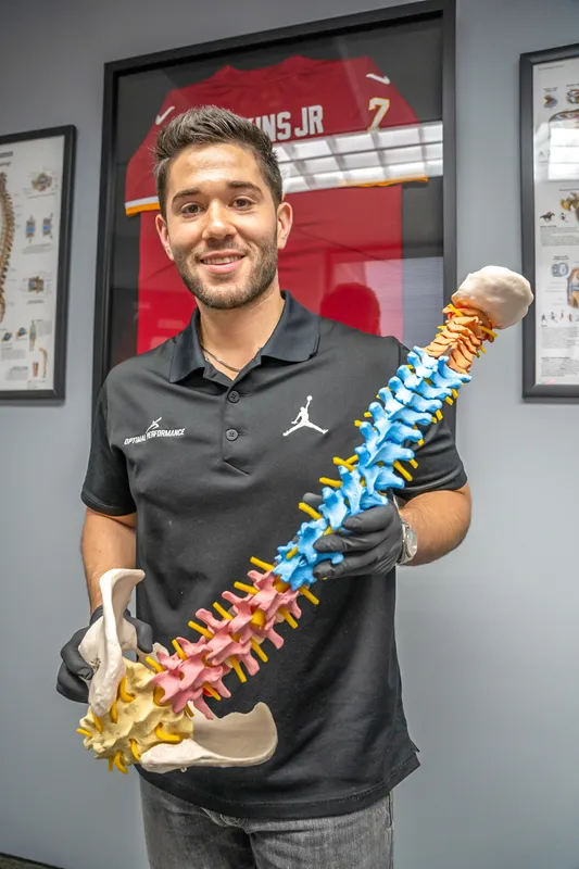 A person wearing a black polo shirt smiles while holding a replica of a human spine. Multiple framed images are on a wall in the background.