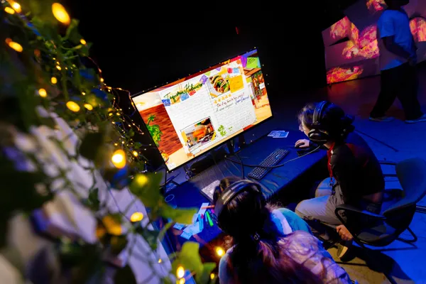 Two people sit in front of a computer screen with headphones on.